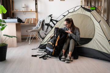 Female Backpacker in eyeglasses and casual clothes sitting into tent with backpack while preparing...