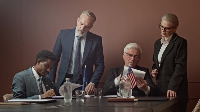 Wide shot of group of diverse representatives and attache examining and signing business contracts sitting at table during intergovernmental meeting