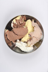 A silver bowl is filled with various scoops of ice cream, including chocolate, vanilla, and mixed flavors, set against a white background