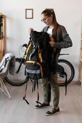 Female Backpacker in shirt and eyeglasses packing backpack with gear for her trip in the living...