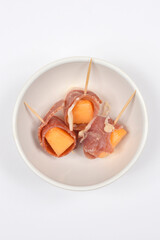 A close-up overhead shot displays three appetizer skewers, each featuring a cube of orange melon wrapped in prosciutto, served in a small white bowl
