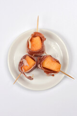 Three elegant appetizers featuring orange melon cubes wrapped in prosciutto and skewered with toothpicks are presented on a white plate