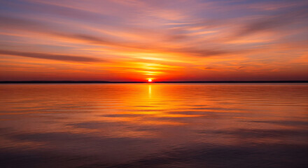 Naklejka premium A breathtaking sunset paints the sky with fiery oranges and purples, reflecting vibrant hues across tranquil water