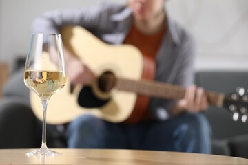 Glass with white wine on table and man playing guitar indoors, selective focus