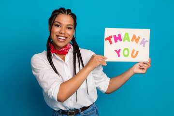 Cheerful young woman holding colorful thank you sign while posing against a bright blue background...
