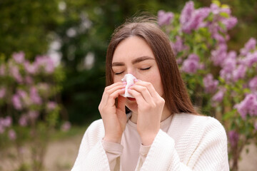 Woman with paper tissue suffering from seasonal pollen allergy near blossoming shrub on spring day,...