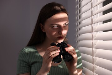 Paranoia. Worried woman with binoculars near window indoors