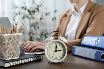 Deadline. Woman with laptop at wooden table in office, focus on alarm clock