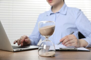 Deadline. Woman with laptop and clipboard working at wooden table in office, focus on hourglass