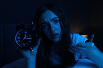 Frustrated woman with alarm clock suffering from insomnia on bed at home