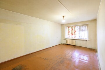 plain room with worn brown floors, off-white walls, a window with patterned curtains, and a radiator. The space is lit by a single ceiling bulb