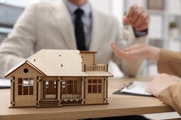 Real estate agent giving key to client at desk in office, focus on house model