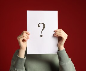 Anonymity. Woman hiding behind paper sheet with question mark on dark red background
