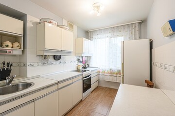 well-lit kitchen with beige cabinets, white countertops, and a wood-look floor. A window with sheer...