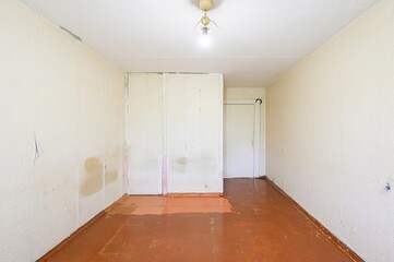 Empty, rundown room with textured walls, bare bulb light, and stained reddish floor. Appears to...
