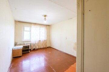 empty room shows peeling wallpaper, a window with lace curtains, a light fixture, and a wood floor