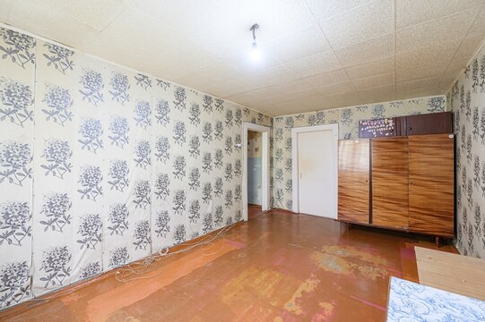 room with floral wallpaper, wooden floor, and a wardrobe. Wires are tangled on the floor. The room looks outdated
