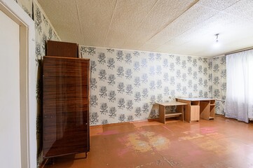 dated room features floral wallpaper, a wooden wardrobe, tables, and worn floors