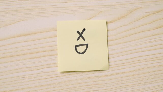 A yellow sticky note with a hand-drawn XD face representing laughter, placed on a wooden surface with a visible hand holding a black marker.