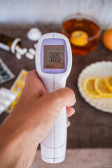 Digital Thermometer Showing 38.6°C in Hand with Medicine and Tea on Nightstand in Background