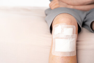 Close-up view of a man's bandaged knee with adhesive patch after injury, sitting on bed. Concept of...