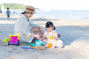 Cute Asian mother and little daughter are sitting on the sand by the sea in the tropical sea of...