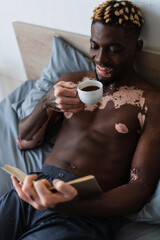 Shirtless african american man with vitiligo smiling while holding coffee and reading book