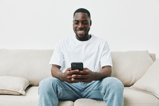 A smiling Black man enjoying social media on his smartphone while sitting on a cozy beige couch, wearing a casual white t shirt and light blue jeans, depicting relaxation and leisure