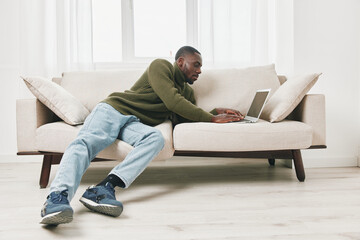 Young Black man working from home on a laptop while lounging on a stylish beige sofa, wearing a...