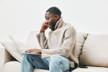 Young African man in a cozy sweater looking surprised while using laptop on a white couch at home,...