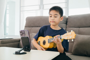 A young boy plays a ukulele on the couch with a smartphone mounted in front for online music...