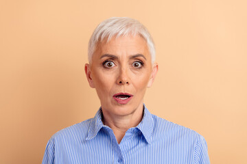 Surprised mature woman with short white hair wearing a blue striped shirt against a beige...