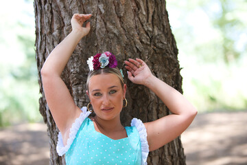 A beautiful young Spanish woman dressed in a traditional green flamenco dress and with a flower in her hair rests leaning against a pine tree at a typical pilgrimage in Andalusia.