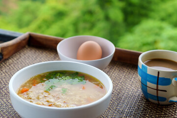 Rice porridge or boiled rice soup with seasoned minced pork, soft -boiled egg in whiite bowl and cup of coffee on wooden tray with green background. Freshy breakfast.