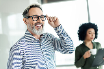 Mature professional man smiling in modern office setting, colleague in background reviewing notes,...