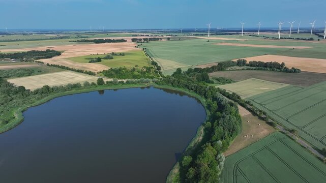 Aerial view of a serene lake reflecting the sky, bordered by lush trees and vibrant fields with distant wind turbines, Penkun, Uckermark, Brandenburg, Germany.
