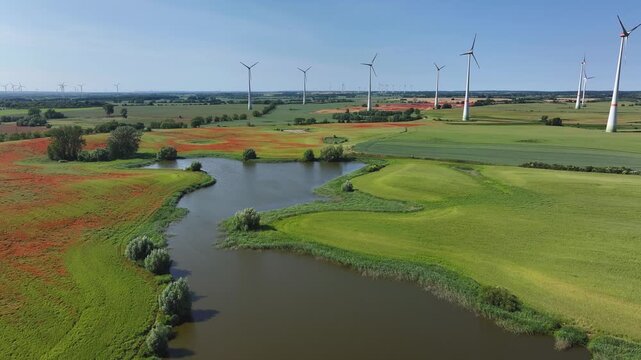 Aerial view of vibrant red poppy fields and wind turbines contrasting with lush green fields and tranquil water, Brussow Uckermark, Brandenburg, Germany.