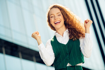 Cheerful businesswoman with curly red hair in green formal wear smiling outdoors against modern...