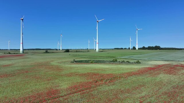 Aerial view of wind turbines standing tall amidst a field dotted with vibrant red poppies against a clear blue sky, Brussow Uckermark, Brandenburg, Germany.