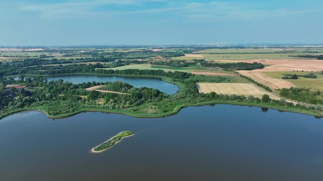 Aerial view of a serene lake contrasted by the vibrant greens of shoreline vegetation and distant wind turbines, Brussow Uckermark, Brandenburg, Germany.