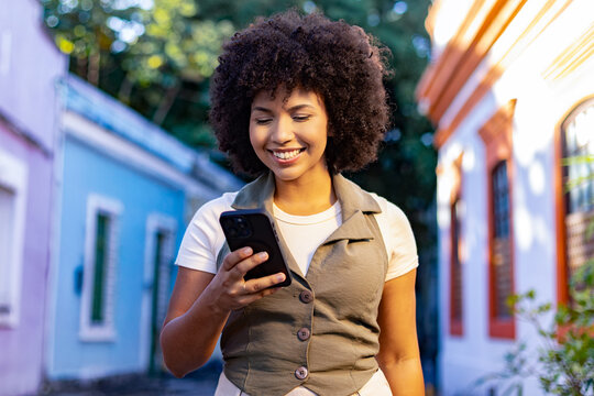 Smiling Afro-Brazilian woman using smartphone outdoors on a colorful street&mdash;modern, connected, and joyful travel lifestyle in Latin America.