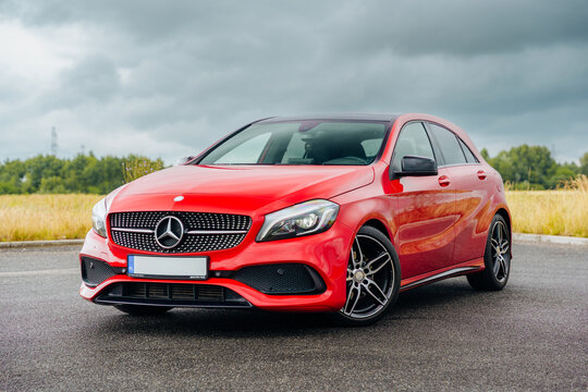 Front view of red Mercedes-Benz A-Class hatchback parked wet asphalt with cloudy sky and countryside. Sporty compact car design captured under natural light for automotive advertising press marketig.