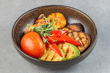 A rustic bowl of vibrant grilled vegetables including zucchini, eggplant, tomato, corn, and red pepper, garnished with fresh greens on a stone surface
