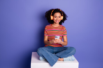 Cheerful teen girl wearing orange headphones holding a smartphone seated on white block against...