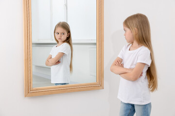 Upset little girl looking at herself in mirror indoors