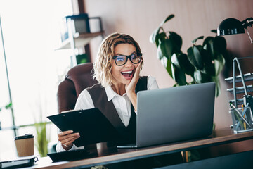 Young businesswoman in a modern office interior working on a laptop and holding a clipboard,...