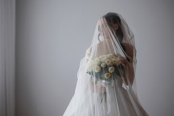 Bride with bouquet stands gracefully behind a veil in softly lit room