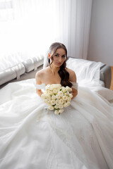Bride in beautiful white gown holds bouquet of roses while sitting in bright room