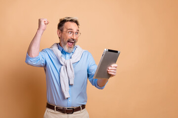 Mature businessman celebrating success while using a tablet, standing against beige background,...