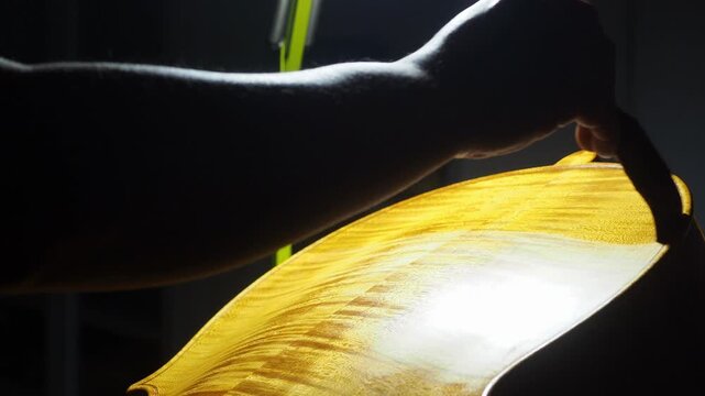 Skilled luthier brushing varnish onto cello back plate marbling, preserving traditional craftsmanship in wooden workshop setting, slow motion